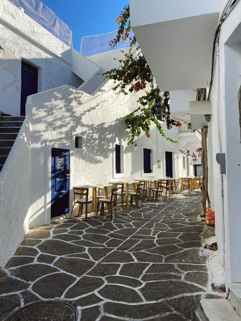 A narrow whitewashed street in Paros with blue doors and outdoor cafe tables under morning sunlight, a top highlight for the best time to visit Greece 2026.