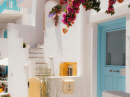 A picturesque Naxos Greece 2026 alley featuring vibrant pink bougainvillea, traditional whitewashed architecture, a blue door, and yellow shutters.