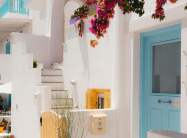 A picturesque Naxos Greece 2026 alley featuring vibrant pink bougainvillea, traditional whitewashed architecture, a blue door, and yellow shutters.