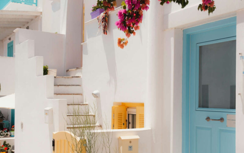 A picturesque Naxos Greece 2026 alley featuring vibrant pink bougainvillea, traditional whitewashed architecture, a blue door, and yellow shutters.