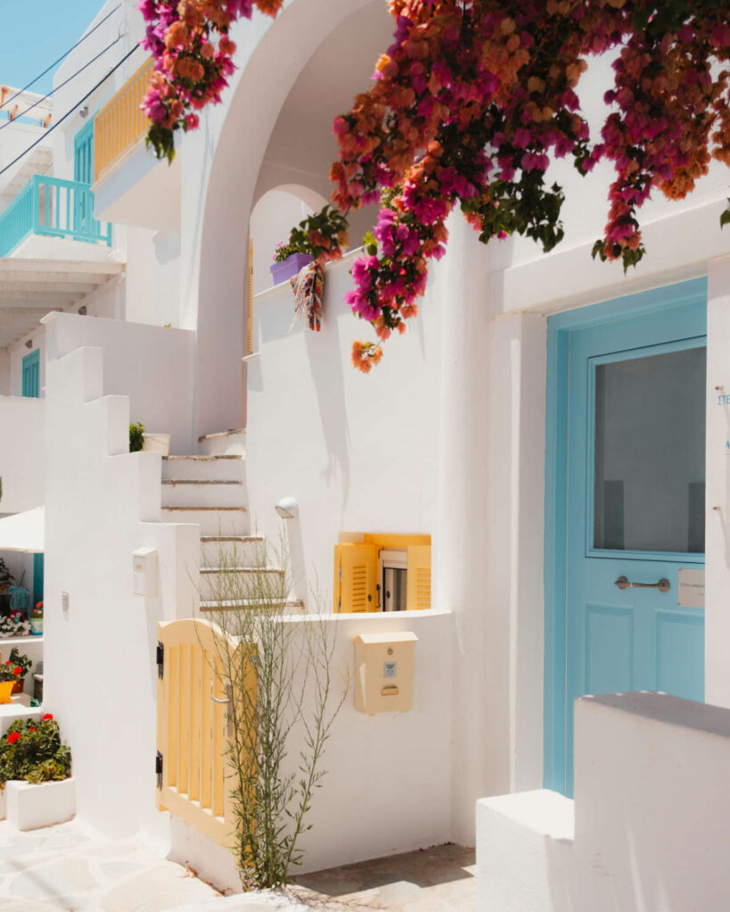 A picturesque Naxos Greece 2026 alley featuring vibrant pink bougainvillea, traditional whitewashed architecture, a blue door, and yellow shutters.