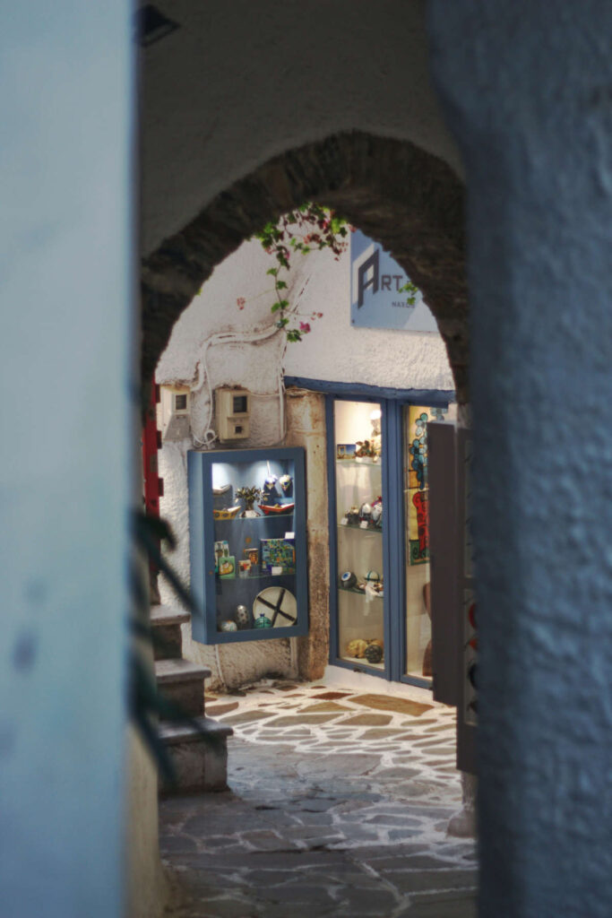 A glimpse through a stone archway into a narrow, traditional Naxos Greece 2026 alley, featuring whitewashed walls, colorful bougainvillea, and a boutique shop with handcrafted items.