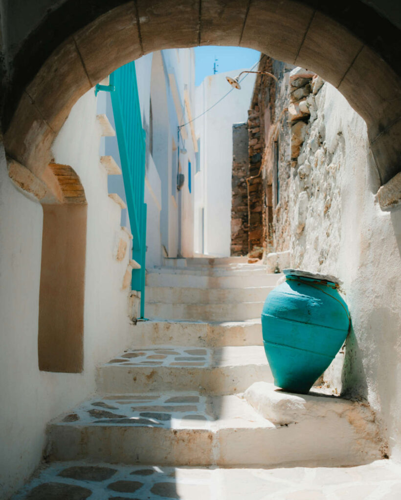 A traditional narrow street in Naxos Greece 2026 featuring whitewashed stone steps, a turquoise pottery jar, and a classic Cycladic archway.