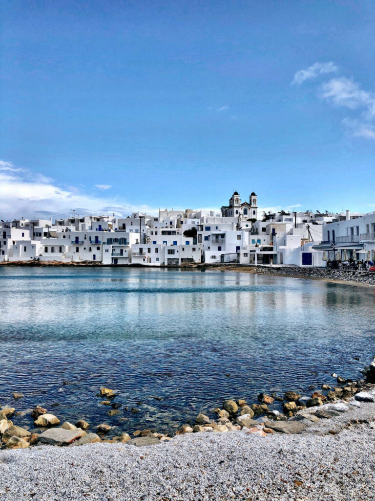 A panoramic view of the traditional whitewashed harbor of Naoussa, Paros Greece 2026, with colorful fishing boats in the turquoise water under a blue sky, perfect for a chic 10-day itinerary.